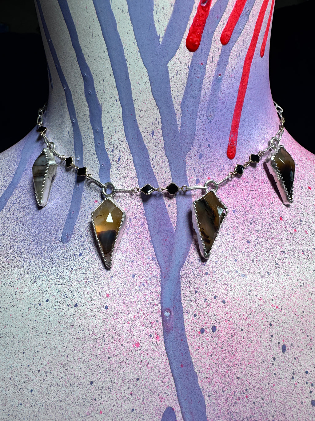 A silver-toned statement necklace with four large faceted brown and amber gemstone pendants and small black beads, displayed on a painted mannequin torso with purple, pink, and red paint drips.
