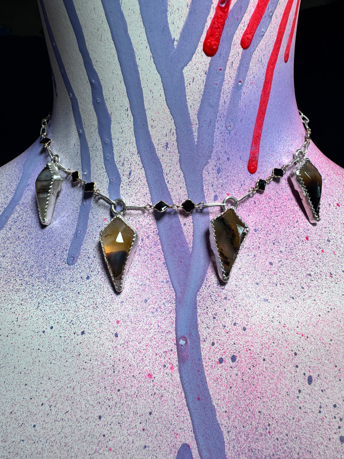 A silver-toned statement necklace with four large faceted brown and amber gemstone pendants and small black beads, displayed on a painted mannequin torso with purple, pink, and red paint drips.