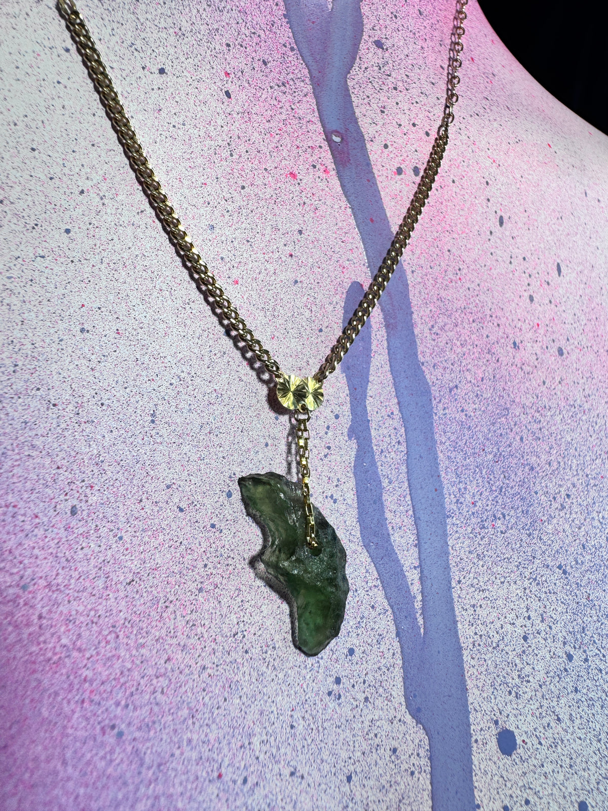 A gold-tone chain necklace with a moldavite charm and a green polished stone pendant, displayed on a textured pink and purple background.