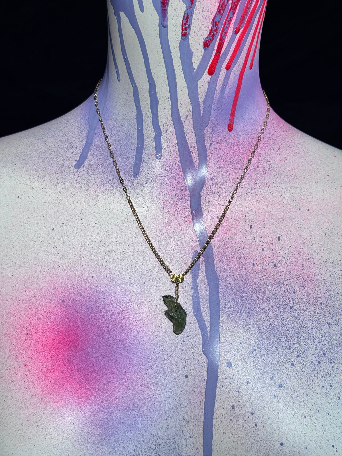 A gold chain necklace with a dark green moldavite pendant displayed on a painted mannequin torso against a black background.