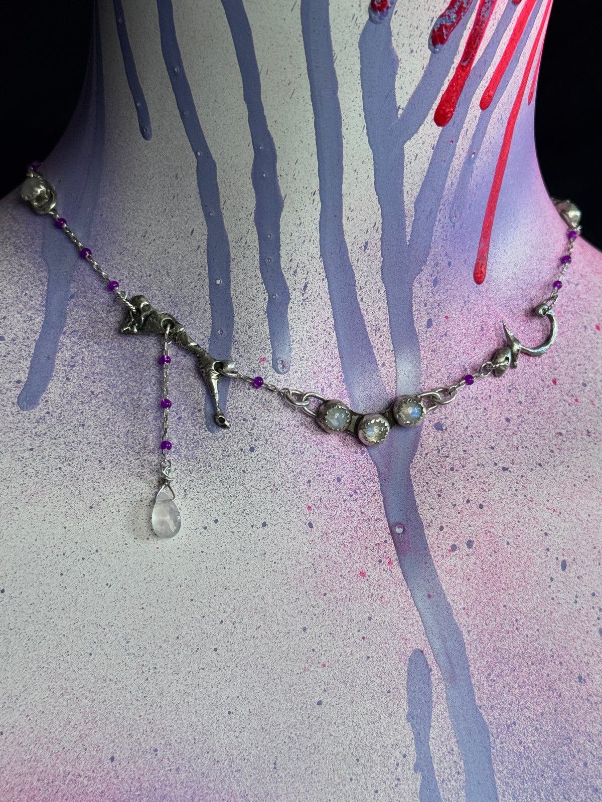 A silver-toned gothic-style necklace with animal charms, purple beads, and a teardrop crystal pendant, displayed on a painted mannequin neck with spray-paint drip details.