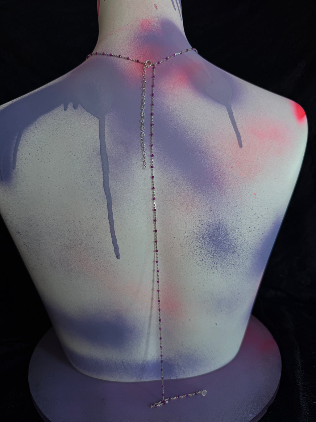 A delicate silver chain necklace with a central drop chain and a small cross pendant, displayed on the back of a spray-painted mannequin torso with purple and pink paint drips against a black background.