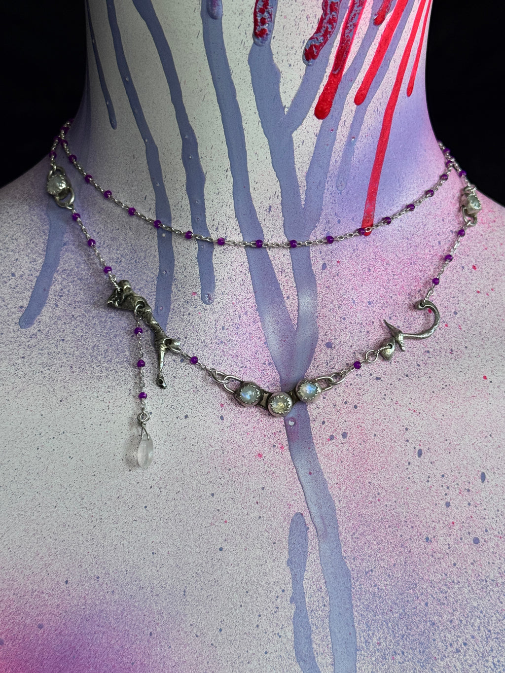A layered silver-toned necklace displayed on a painted mannequin bust. The necklace features silver-toned metal links, small purple beads, iridescent oval stones, and a dangling clear faceted bead.