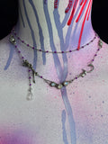 A layered silver-toned necklace displayed on a painted mannequin bust. The necklace features silver-toned metal links, small purple beads, iridescent oval stones, and a dangling clear faceted bead.