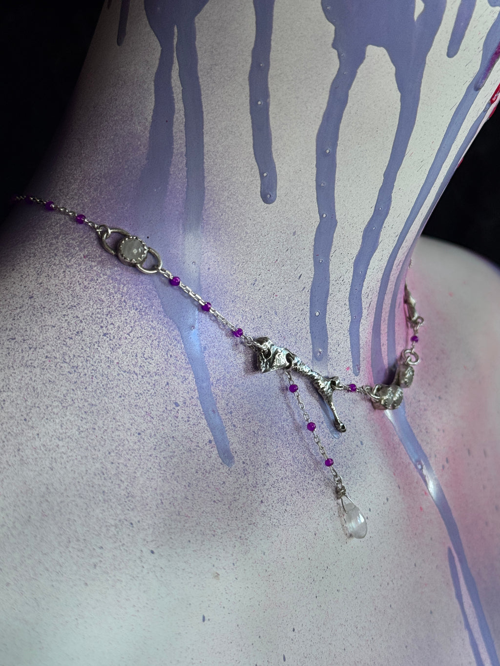 A silver-toned beaded necklace with small purple beads, a silver cross pendant, and a dangling clear teardrop crystal, displayed on a painted neck with purple drips and pink shading.