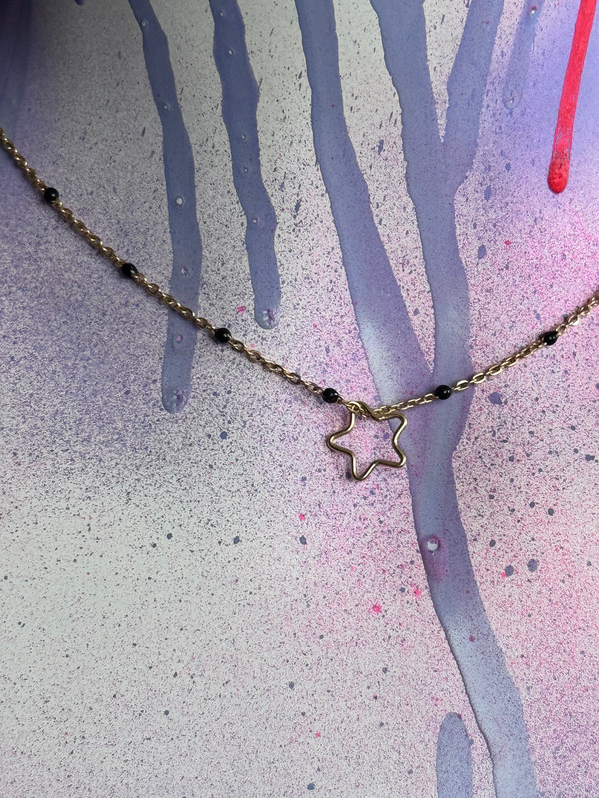 A delicate gold chain necklace with a small star-shaped pendant and tiny dark beads, displayed against a textured, paint-splattered background.