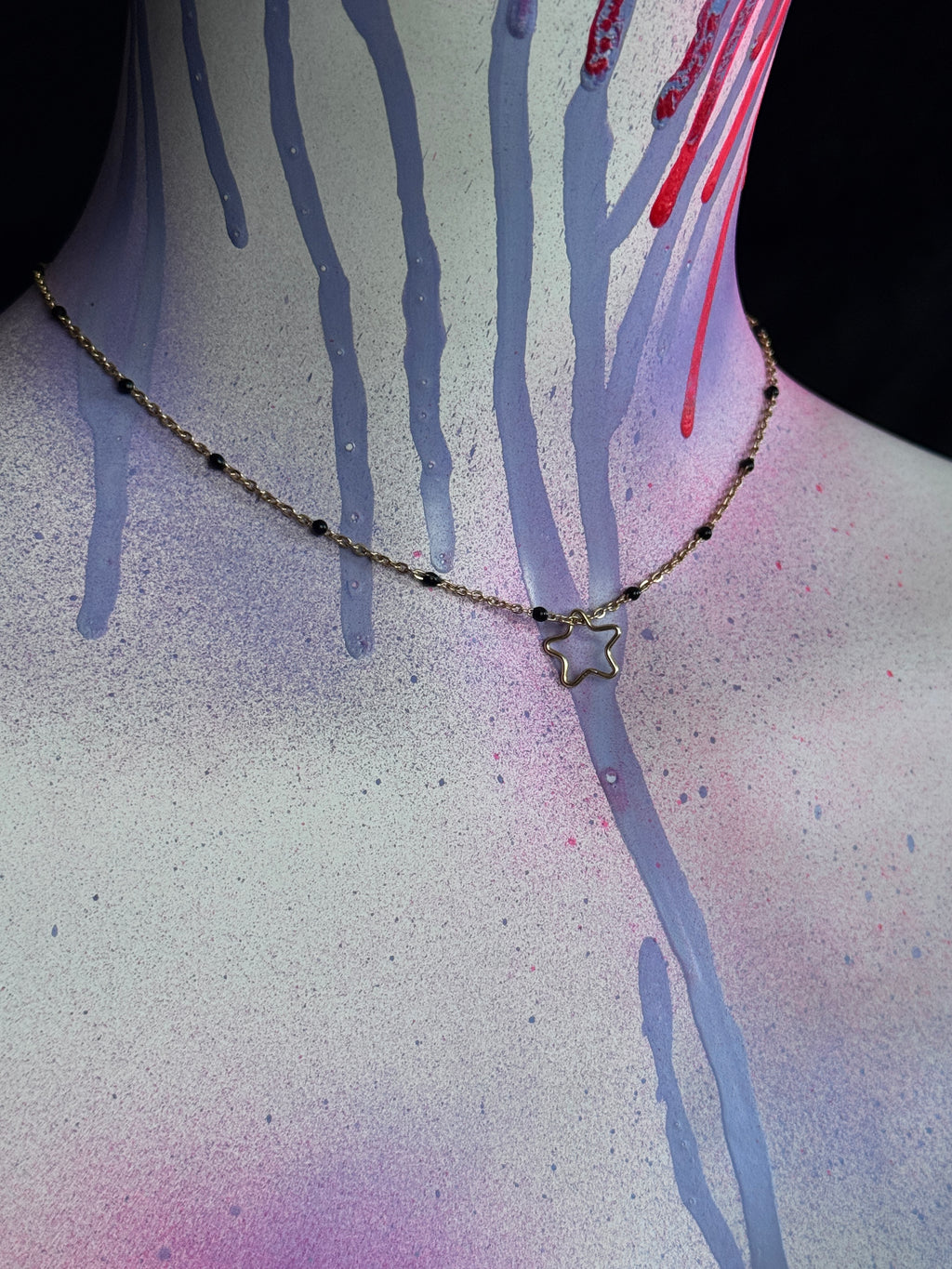 A delicate gold-toned chain necklace with a small star-shaped pendant, shown on a mannequin or model neck against a dark background with spray-paint drips in purple, pink, and red.