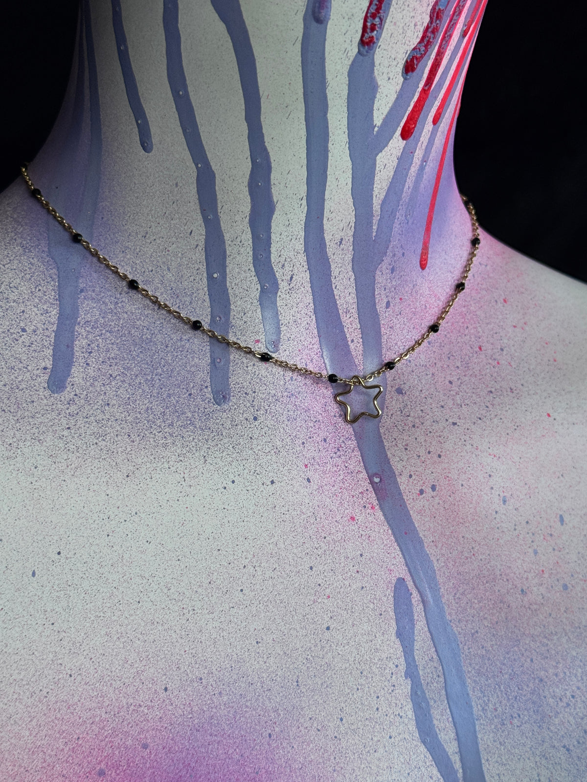A delicate gold-toned chain necklace with a small star-shaped pendant, shown on a mannequin or model neck against a dark background with spray-paint drips in purple, pink, and red.