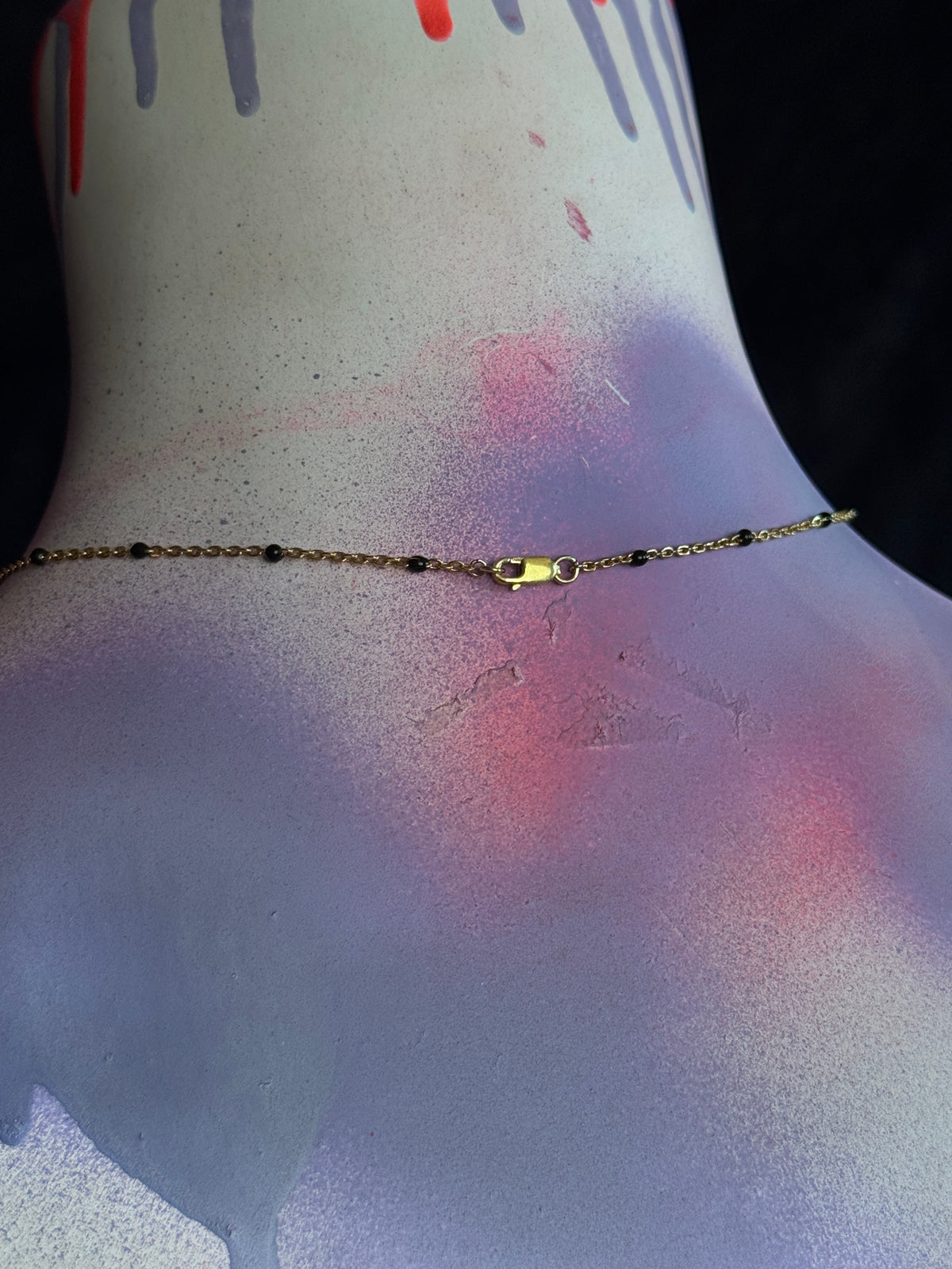 A delicate gold chain anklet with small dark beads and a lobster clasp, shown worn on the ankle of a mannequin or model with a painted, textured surface.