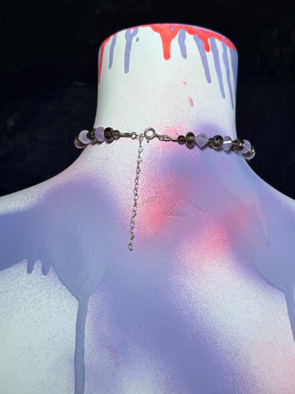 A beaded choker necklace with dark brown beads and lavender-purple heart-shaped beads, displayed on a white mannequin bust with spray-paint drip details under dramatic purple and pink lighting.