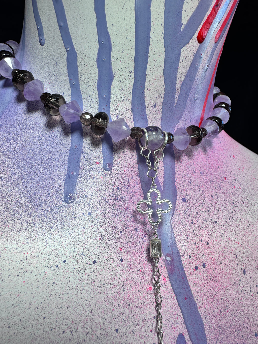 A beaded necklace with faceted purple and smoky black beads, a silver-toned cross charm, and a dangling clear rectangular bead, displayed on a painted mannequin bust with purple, pink, and blue paint drips.