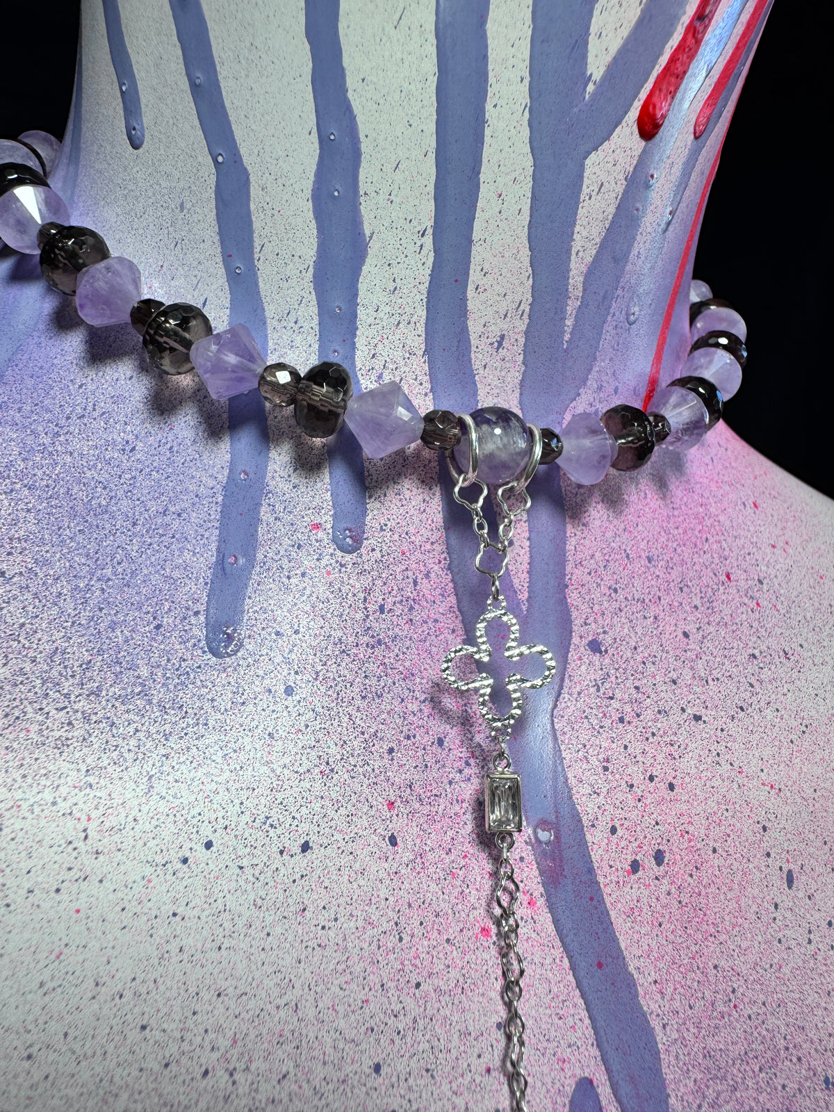 A beaded necklace with faceted purple and smoky black beads, a silver-toned cross charm, and a dangling clear rectangular bead, displayed on a painted mannequin bust with purple, pink, and blue paint drips.