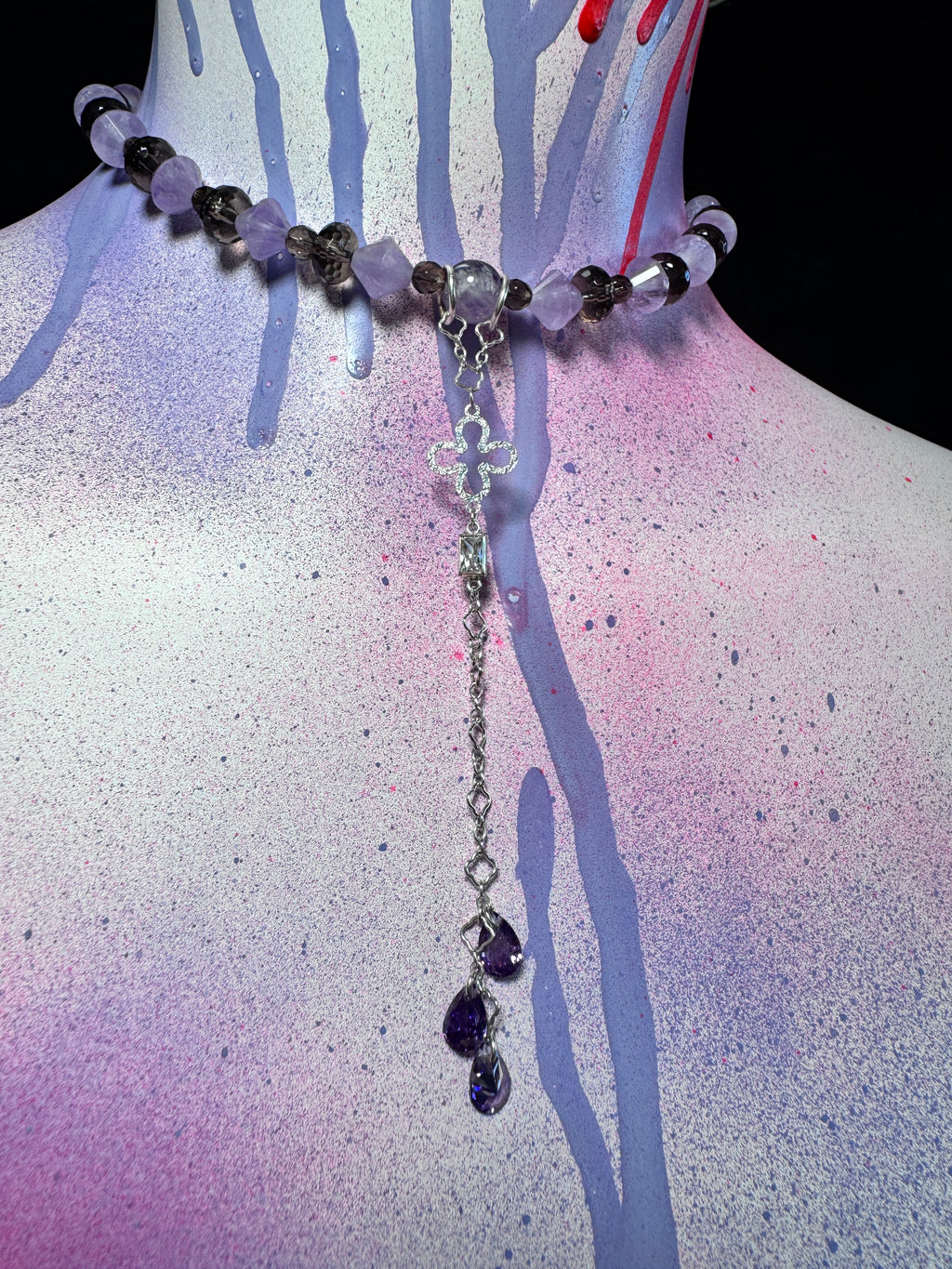 A beaded necklace displayed on a painted mannequin bust. The necklace features light purple and translucent white beads, dark gray beads, a silver-toned cross centerpiece, and two dangling purple teardrop crystals at the bottom.