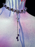 A beaded necklace displayed on a painted mannequin bust. The necklace features light purple and translucent white beads, dark gray beads, a silver-toned cross centerpiece, and two dangling purple teardrop crystals at the bottom.