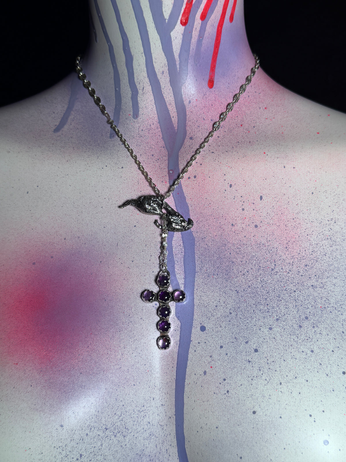 A gothic-style silver-toned necklace featuring a bird and silver pendant with amethyst gemstone accents, displayed on a mannequin torso decorated with spray-paint splatters and drips.