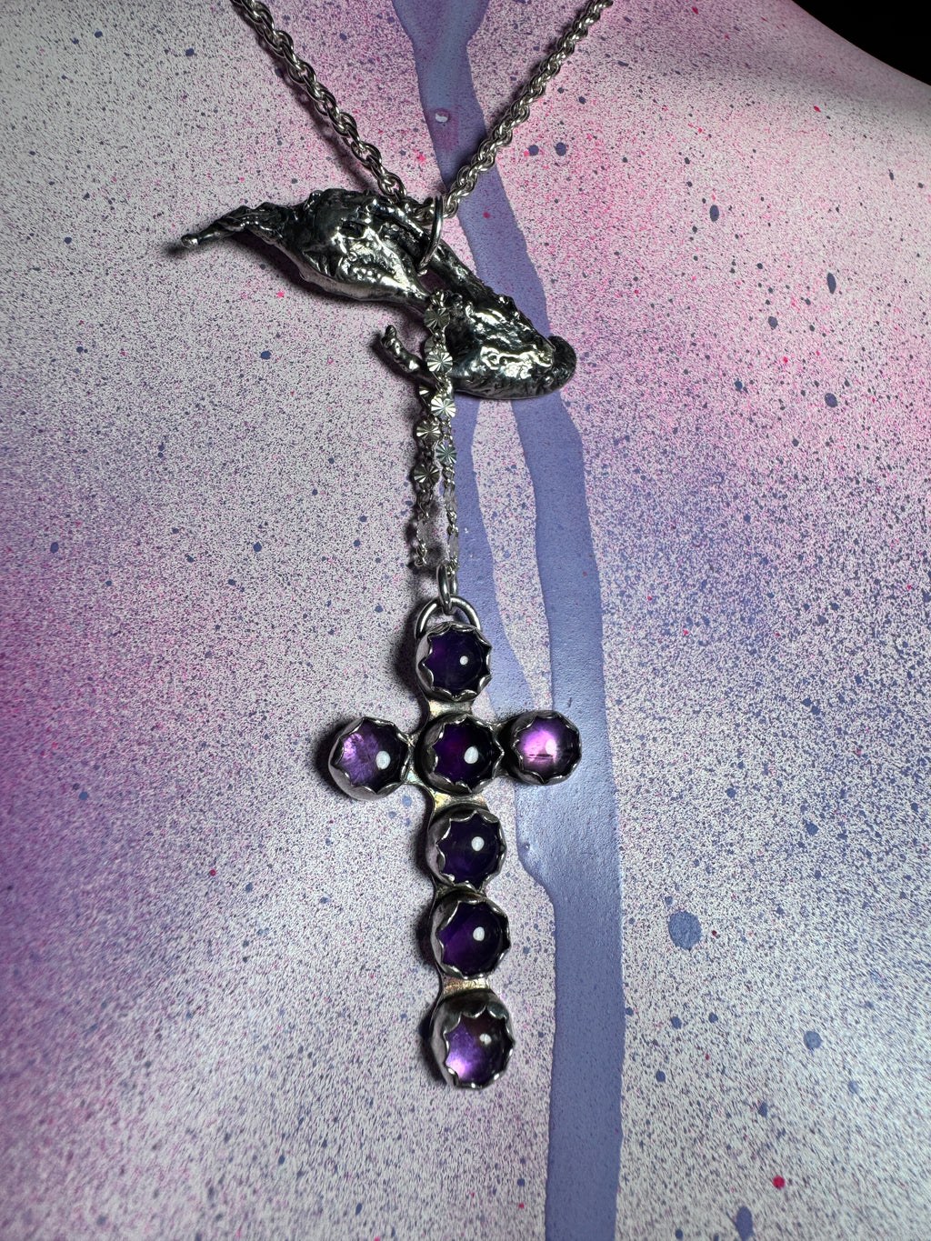 A silver-toned cross pendant necklace featuring five faceted amethyst gemstones, suspended from a matching chain with a textured silver charm, displayed on a pink and purple paint-splattered surface.