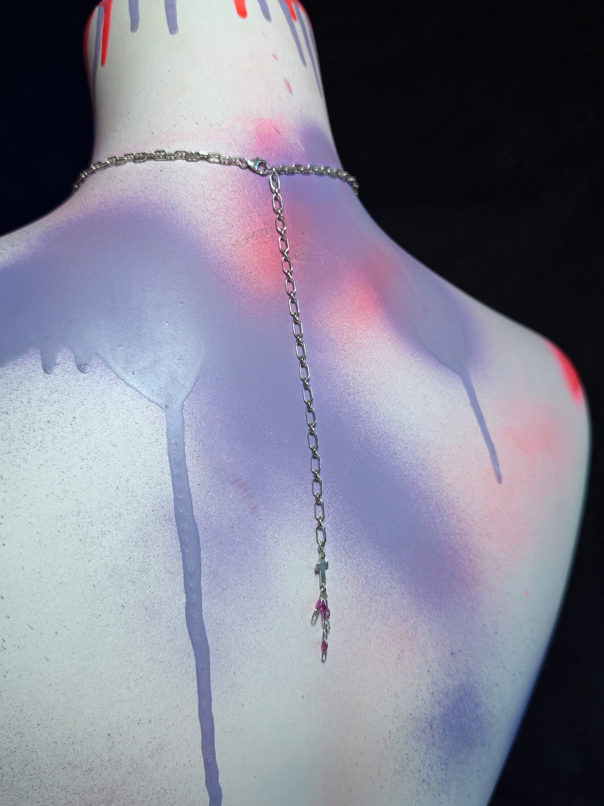 A silver-toned chain necklace with a long vertical drop and small amethyst gemstone accents, displayed on the back of a painted mannequin torso against a black background.