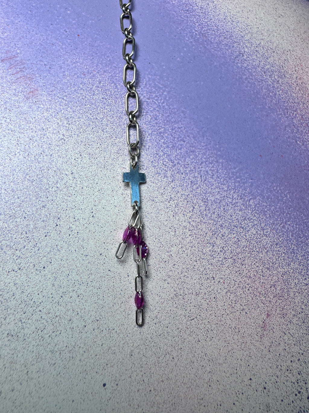 A silver-toned metal chain necklace with a small cross pendant, silver paperclip charms, and purple beads, displayed on a textured purple and blue background.
