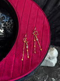 A pair of gold-toned dangle earrings displayed on a deep magenta velvet jewelry stand, with a dark textured background and a small white stone prop.