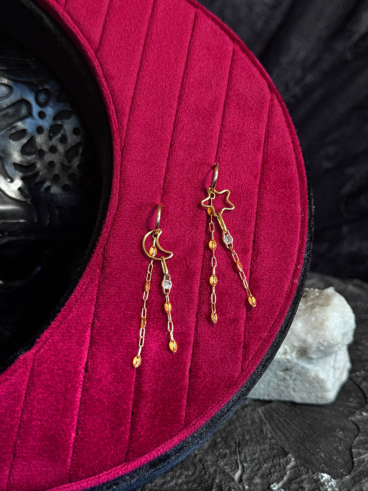 A pair of gold-toned dangle earrings displayed on a deep magenta velvet jewelry stand, with a dark textured background and a small white stone prop.