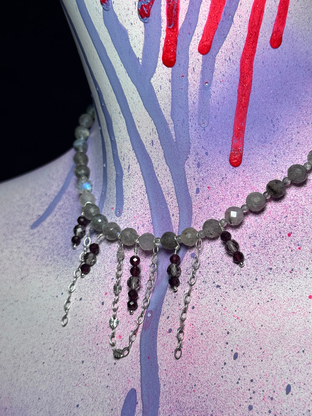A beaded necklace with faceted gray and dark purple beads, featuring silver-toned chains and dangling charms, displayed on a painted mannequin torso with purple, pink, and red paint splatters.
