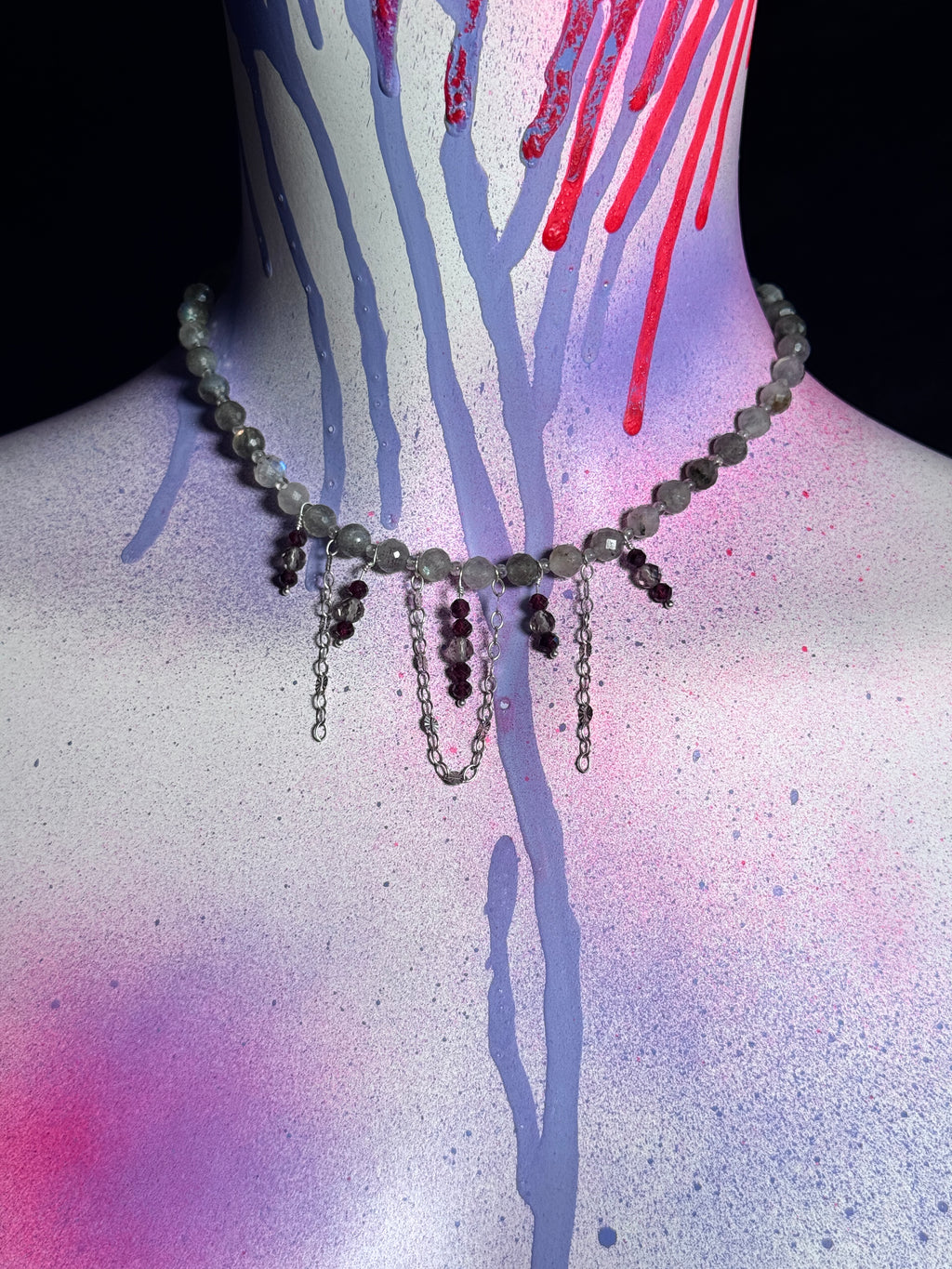 A beaded choker necklace with dangling chains and faceted beads, displayed on a mannequin neck against a black background with colorful paint drips.