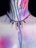 A beaded choker necklace with dangling chains and faceted beads, displayed on a mannequin neck against a black background with colorful paint drips.