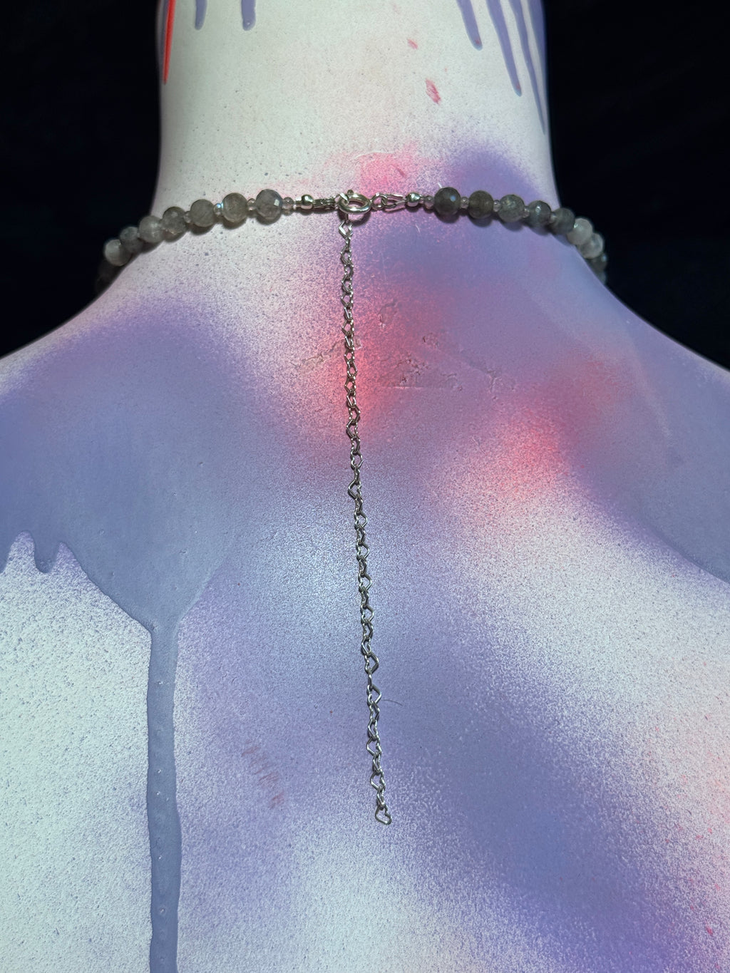 A beaded choker necklace with a long dangling chain, displayed on a painted mannequin torso. The beads are a muted grayish-green, and the metal chain and clasp appear silver-toned.